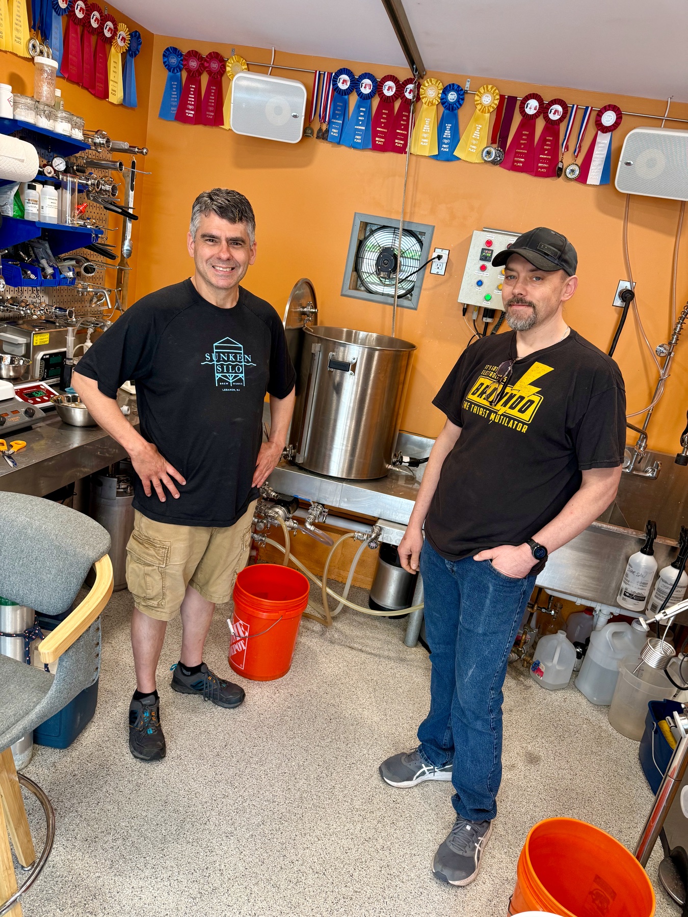 Two brewers in the orange garage brewery beneath competition ribbons